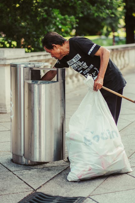 A man with dark hair, wearing a black T-shirt with white text and dark trousers, is seen on a paved outdoor pathway, leaning over a cylindrical stainless steel public waste bin to dispose of litter. He holds a large white trash bag with both hands, which appears to be filled with waste, and is in the process of emptying it into the bin. The bin is situated near a low stone balustrade, with lush green foliage and trees visible in the background, suggesting a park or landscaped urban area. The scene is well-lit with natural daylight, highlighting the metallic surface of the waste bin, the man's focused posture, and the texture of the trash bag. This image visually relates to waste collection and rubbish disposal, emphasizing urban cleanliness and private waste handling services provided by Waste Removal Fulham, supporting the theme of alternative rubbish removal options in a public environment.