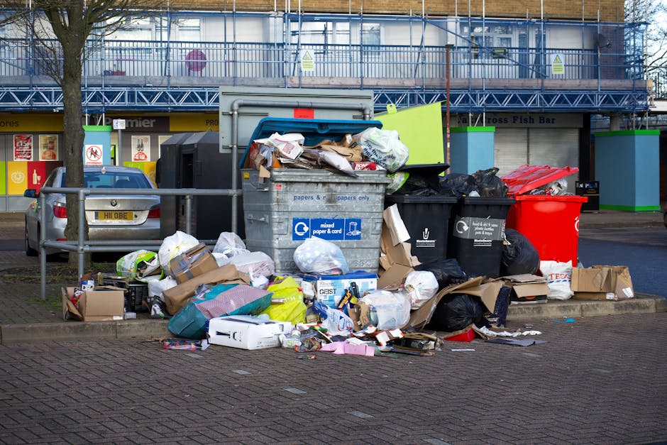 A collection of overflowing rubbish bins and assorted waste materials is situated on an urban pavement near a parking area. The scene includes a large metal bin labeled for mixed paper and cardboard, which is partially open with shredded paper, cardboard boxes, and plastic waste visible inside. Surrounding the bin are black trash bags, some torn open, revealing crumpled paper, plastic bottles, and packaging materials. To the right, a red recycling bin and a black bin are also placed, with additional loose waste scattered on the ground, including flattened cardboard boxes, plastic wrappers, and paper debris. Behind the waste collection point, there is a street with a parked car, and a commercial building with storefronts and a blue metal fire escape staircase attached to the facade. The environment appears to be an outdoor public space, with a tree and a metal railing in front of the waste area, illustrating an example of off-site waste accumulation that waste removal services such as Waste Removal Fulham can handle through private disposal solutions, avoiding reliance on local authority collection.