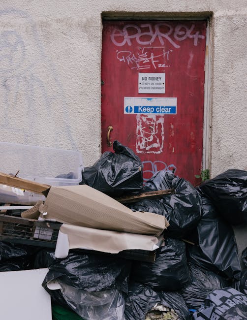 A collection of black rubbish bags filled with waste, piled up against an exterior wall beneath a red metal door that has graffiti, stickers, and signs attached. The bags are made of plastic, some torn open or bulging, with a few cardboard and paper waste visible among them. The door is weathered with peeling paint, graffiti tags in white and pink spray paint, and stickers including one with the message 'Keep clear' and another indicating 'No money.' The surrounding wall appears textured with a beige, slightly rough surface, contrasting with the smoother finishes of the bags and debris. The environment suggests a back alley or service access point where private waste collection might occur, emphasizing the context of independent rubbish disposal. Waste Removal Fulham’s service context is subtly implied by the scene of unmanaged waste outside a typical urban property entrance, underscoring the importance of proper rubbish removal and the potential need for professional onsite clearance in Fulham flats.