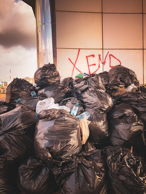 A collection of black rubbish bags filled with waste, piled up against an exterior wall beneath a red metal door that has graffiti, stickers, and signs attached. The bags are made of plastic, some torn open or bulging, with a few cardboard and paper waste visible among them. The door is weathered with peeling paint, graffiti tags in white and pink spray paint, and stickers including one with the message 'Keep clear' and another indicating 'No money.' The surrounding wall appears textured with a beige, slightly rough surface, contrasting with the smoother finishes of the bags and debris. The environment suggests a back alley or service access point where private waste collection might occur, emphasizing the context of independent rubbish disposal. Waste Removal Fulham’s service context is subtly implied by the scene of unmanaged waste outside a typical urban property entrance, underscoring the importance of proper rubbish removal and the potential need for professional onsite clearance in Fulham flats.