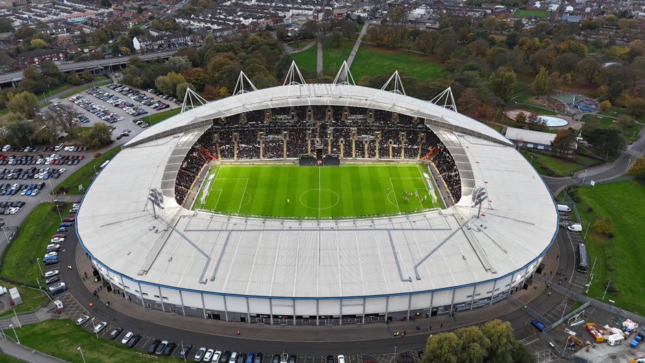 An aerial view of Craven Cottage stadium, showcasing its oval-shaped white roof supported by multiple white masts. The stadium's interior reveals a vividly green grass pitch marked for football, with white lines and goal areas at each end. Surrounding the pitch are dark seating sections filled with spectators, and the exterior of the structure is composed of white and grey materials with some blue accents. The stadium is situated amidst a mix of greenery, including trees and parkland, with adjacent parking lots filled with vehicles on the left side of the image. A roadway encircles the stadium, with some parked cars along the edges and a few service vehicles nearby. The background includes a residential area and scattered trees, with the whole scene bathed in natural daylight under an overcast sky. This scene exemplifies an independent football stadium, often requiring private waste management solutions such as rubbish clearance services to manage waste generated during match days, aligning with services offered by Waste Removal Fulham.