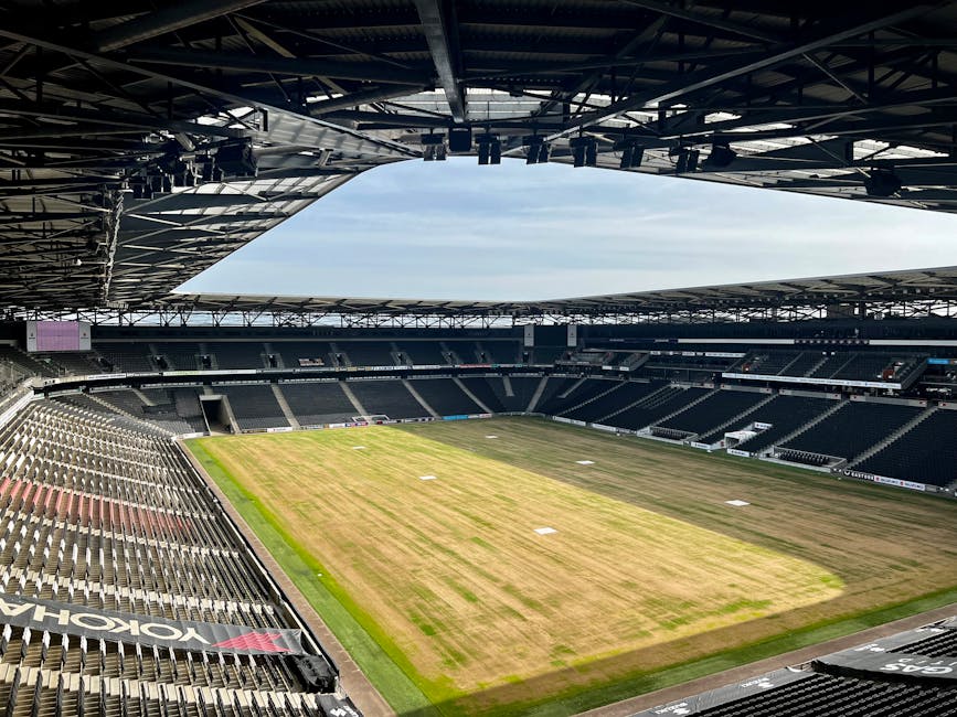 An empty football stadium with a grass pitch that shows signs of recent wear and patchy discoloration, primarily in shades of green and brown. The seating areas surround the pitch on all sides, featuring dark-colored seats arranged in multiple tiers, with some sections appearing more densely packed than others. The stadium roof, constructed from metal framework and supported by large beams, partially covers the seating areas and has a cut-out section allowing natural light to illuminate the field below. The sky visible through this opening is partly cloudy, with a mix of light blue and thin white clouds. The environment is clean and well-maintained, with no visible rubbish or debris. Around the edges of the pitch, zones of lighter and darker grass suggest recent or ongoing maintenance, possibly related to ground management services. This scene exemplifies a professional sports venue prepared for a match, reflecting the importance of regular cleaning and upkeep, which a waste removal company such as Waste Removal Fulham could support through on-site clearance and waste handling services before and after events.