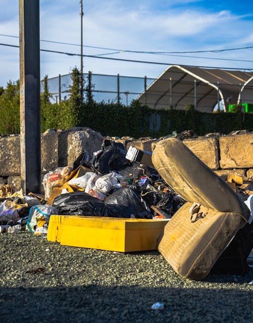 A roadside pile of mixed waste materials is visible in the foreground, including black plastic rubbish bags, a yellow plastic container, and a used, stained car tire lying on its side. Behind these objects, there is a weathered stone wall made of irregular, rough-textured stones stacked without mortar, separating the waste area from a background featuring a chain-link fence, some greenery, and a large outdoor sports or tennis court covered with a curved, semi-transparent canopy supported by metal framework. The scene is illuminated by natural daylight, casting shadows from objects onto the gravel and asphalt surface. Utility poles and overhead power lines cross the sky, which has a few scattered clouds, suggesting a clear day. The image relates to independent waste collection or on-site rubbish clearance, as provided by waste removal services such as Waste Removal Fulham, emphasizing the importance of proper disposal and management of waste materials in urban environments.