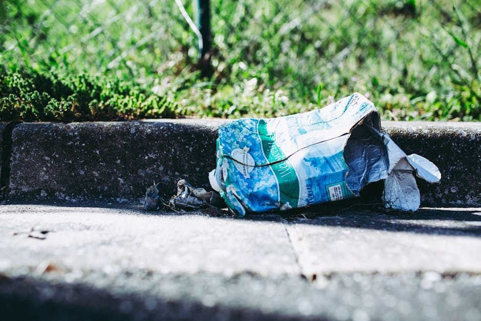 A crumpled plastic bag with a blue and white design lies discarded on a concrete sidewalk next to a low curb, with dirt and debris scattered nearby. The bag appears to be made of thin, glossy plastic material, with some parts torn or wrinkled. In the background, there is a patch of green grass and small plants, indicating an outdoor environment. The scene is illuminated by natural daylight, casting shadows on the pavement. The overall setting suggests a typical urban or suburban street scene where waste materials such as litter may be found, highlighting the context for rubbish removal services like those offered by Waste Removal Fulham in managing private disposal and non-authority collection efforts.