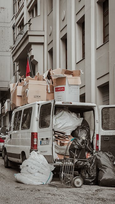 A white panel van parked on a city street beside a beige multi-storey building with tall windows and architectural detailing. The back doors of the van are open, revealing an interior filled with various discarded items, including large black trash bags, flattened cardboard boxes, and a rolled-up white plastic sheet. Several cardboard boxes, some partially crushed or ripped, are stacked on top of the van, with a few resting on the roof, indicating an on-site waste collection or removal process. In front of the van, additional black trash bags and loose recyclables are laid on the pavement, along with a small hand truck and a trolley, suggesting organized waste handling. The scene is illuminated by natural daylight, casting soft shadows and highlighting the mix of packaging materials, textures, and neutral tones. This visual depicts private rubbish collection efforts, aligning with independent waste removal services, such as those offered by Waste Removal Fulham, for effective rubbish clearance near urban residential or commercial premises.