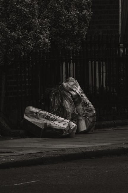 Three large black garbage bags, made of shiny plastic material, are placed on the edge of a paved curb next to a black metal fence and dense foliage. The bags appear to contain waste materials, with one of them slightly torn open near the top. The environment suggests an outdoor urban setting, possibly in a residential area, with natural light casting subtle shadows. The scene reflects a typical instance of rubbish awaiting collection, which could relate to private waste disposal services provided by companies like Waste Removal Fulham. The bags are positioned on a paved sidewalk beside a street, emphasizing the importance of timely rubbish removal to maintain cleanliness and prevent delays in waste collection. The background includes a wooden lattice structure and parts of a typical suburban garden, with dense leaves and dark shadows contributing to the overall muted appearance, consistent with a neutral and factual description suitable for accessibility purposes.