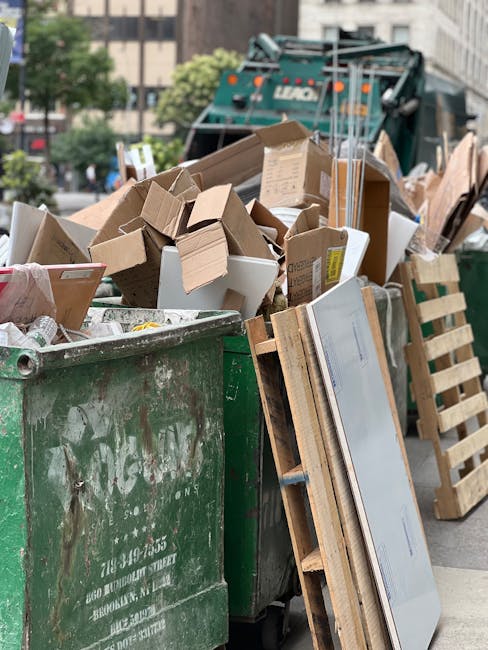 A large green waste disposal bin filled with various types of waste, including corrugated cardboard boxes, some flattened and others partially collapsed, as well as smaller packaging materials, all showing signs of dirt and weathering. The bins are placed on an urban street area with a background that includes a municipal waste collection vehicle, likely used for handling private or alternative rubbish disposal, visible in front of multi-storey buildings and trees. To the right of the bin, there are wooden pallets leaning against the side, and additional cardboard boxes and packaging materials are stacked nearby. The scene is illuminated by natural daylight, with a slightly overcast sky, suggesting daytime city environment. The presence of a waste collection truck and variety of waste materials demonstrates an example of on-site clearance or private waste handling, rather than standard council rubbish collection. The scene exemplifies typical arrangements for independent rubbish removal services, aligning with alternative waste management practices.