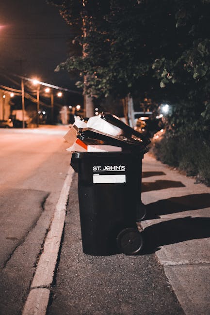 Three large black garbage bags, made of shiny plastic material, are placed on the edge of a paved curb next to a black metal fence and dense foliage. The bags appear to contain waste materials, with one of them slightly torn open near the top. The environment suggests an outdoor urban setting, possibly in a residential area, with natural light casting subtle shadows. The scene reflects a typical instance of rubbish awaiting collection, which could relate to private waste disposal services provided by companies like Waste Removal Fulham. The bags are positioned on a paved sidewalk beside a street, emphasizing the importance of timely rubbish removal to maintain cleanliness and prevent delays in waste collection. The background includes a wooden lattice structure and parts of a typical suburban garden, with dense leaves and dark shadows contributing to the overall muted appearance, consistent with a neutral and factual description suitable for accessibility purposes.